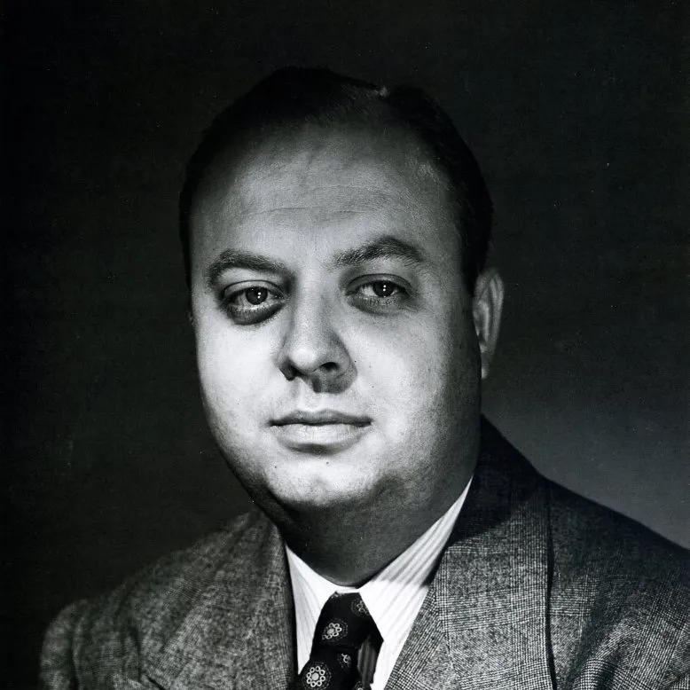 black and white studio portrait of Bruce Bellas in a tweed jacket, striped shirt, and patterned tie, against a dark background.