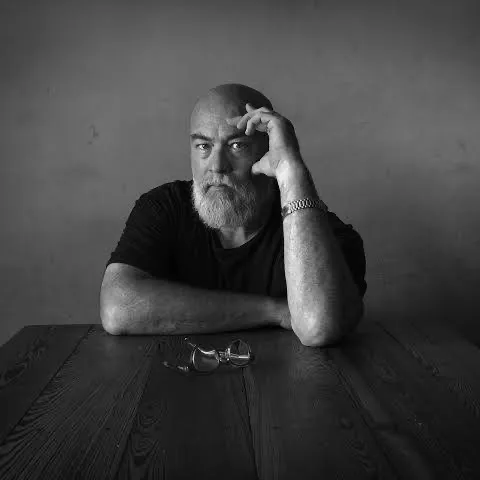 black and white photograph of Dan Winters seated at a wooden table, resting his head on one hand, with a pair of glasses on the table in front of him.