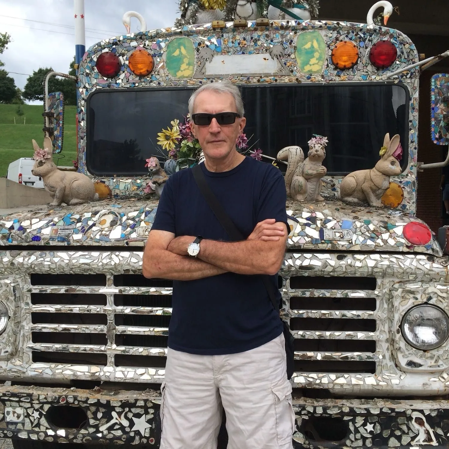color photograph of John Humble with arms crossed standing in front of a mosaic-covered bus decorated with rabbit and squirrel figures.