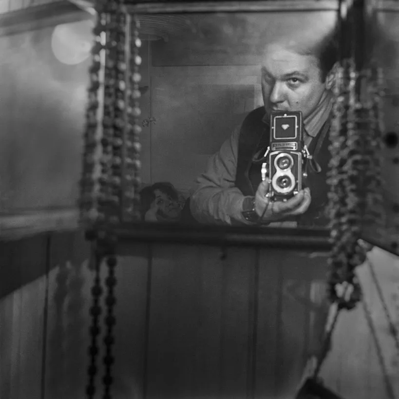 black and white self-portrait of Larry Fink in a mirror framed by hanging beads, holding a twin-lens reflex camera at his chest.