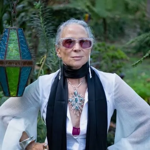 color photograph of Lisa Eisner in jeweled sunglasses, a black scarf, a large turquoise pendant, and a flowing white blouse, standing in a garden beside a stained glass lantern.