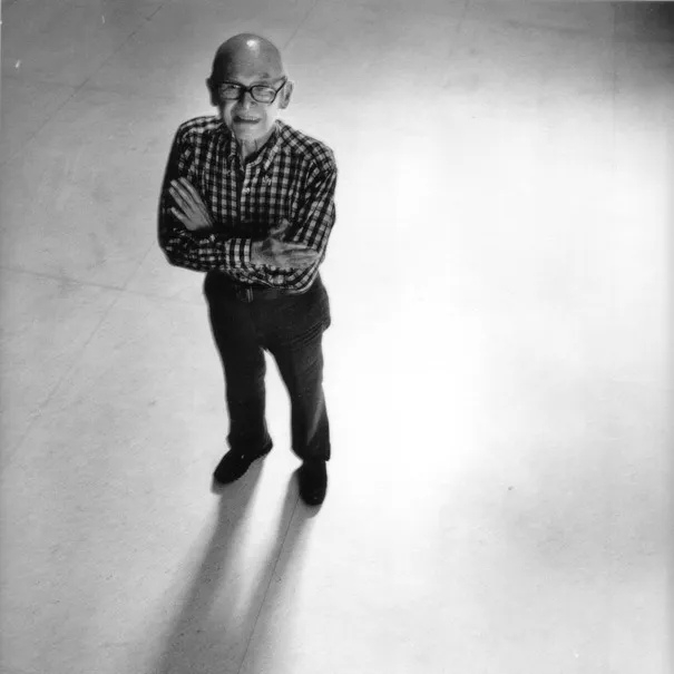 black and white photograph taken from above of Max Yavno in glasses and a checked shirt, arms crossed, standing on a pale floor with his shadow stretching behind him.