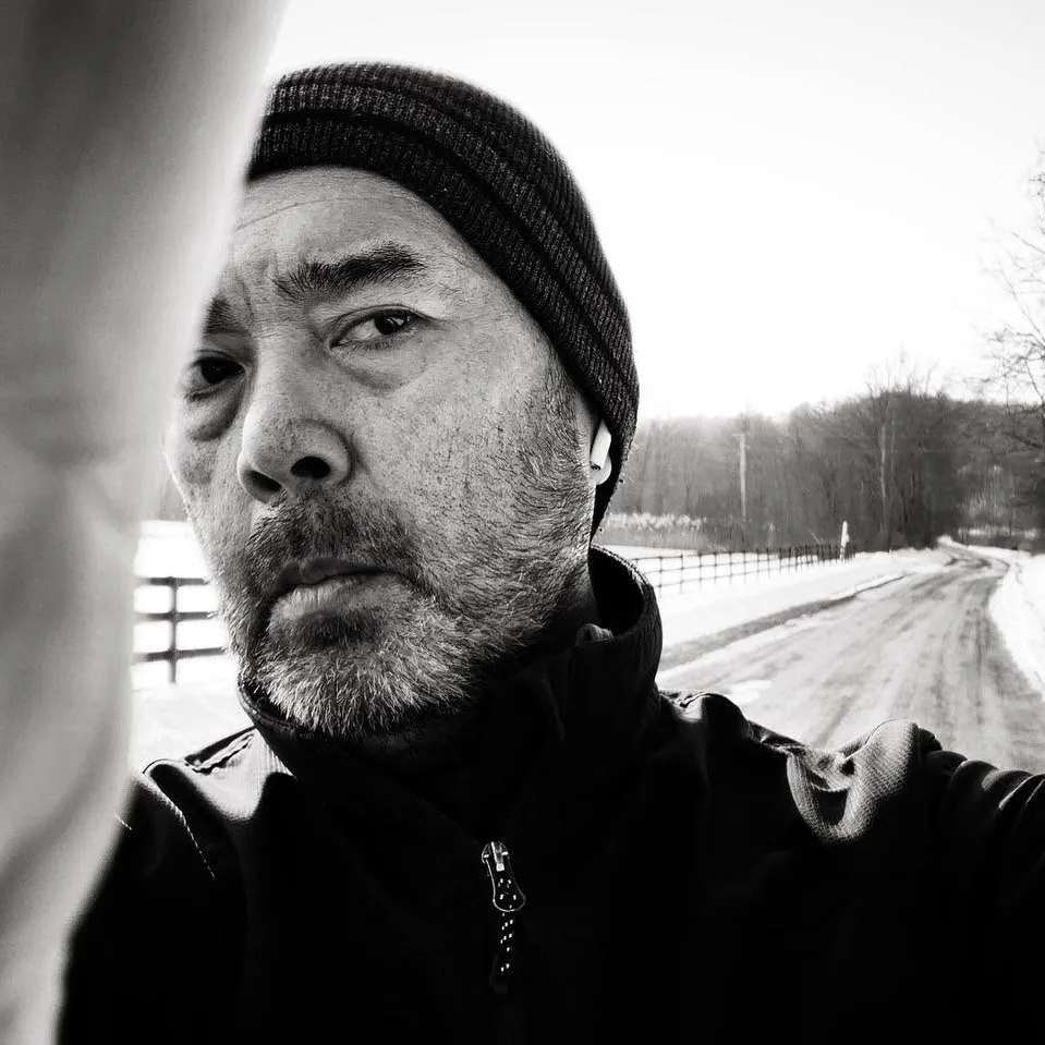 black and white photograph of Michael Lardizabal in a knit cap and dark jacket, looking into the camera on a snowy country road lined with fences and bare trees.