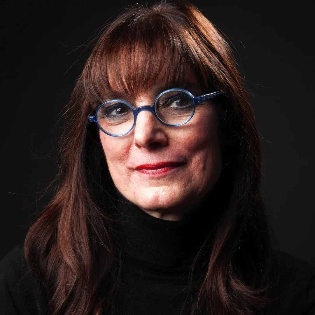color photograph of Nancy Burson in round blue glasses and a black turtleneck, with long auburn hair and bangs, against a black background.