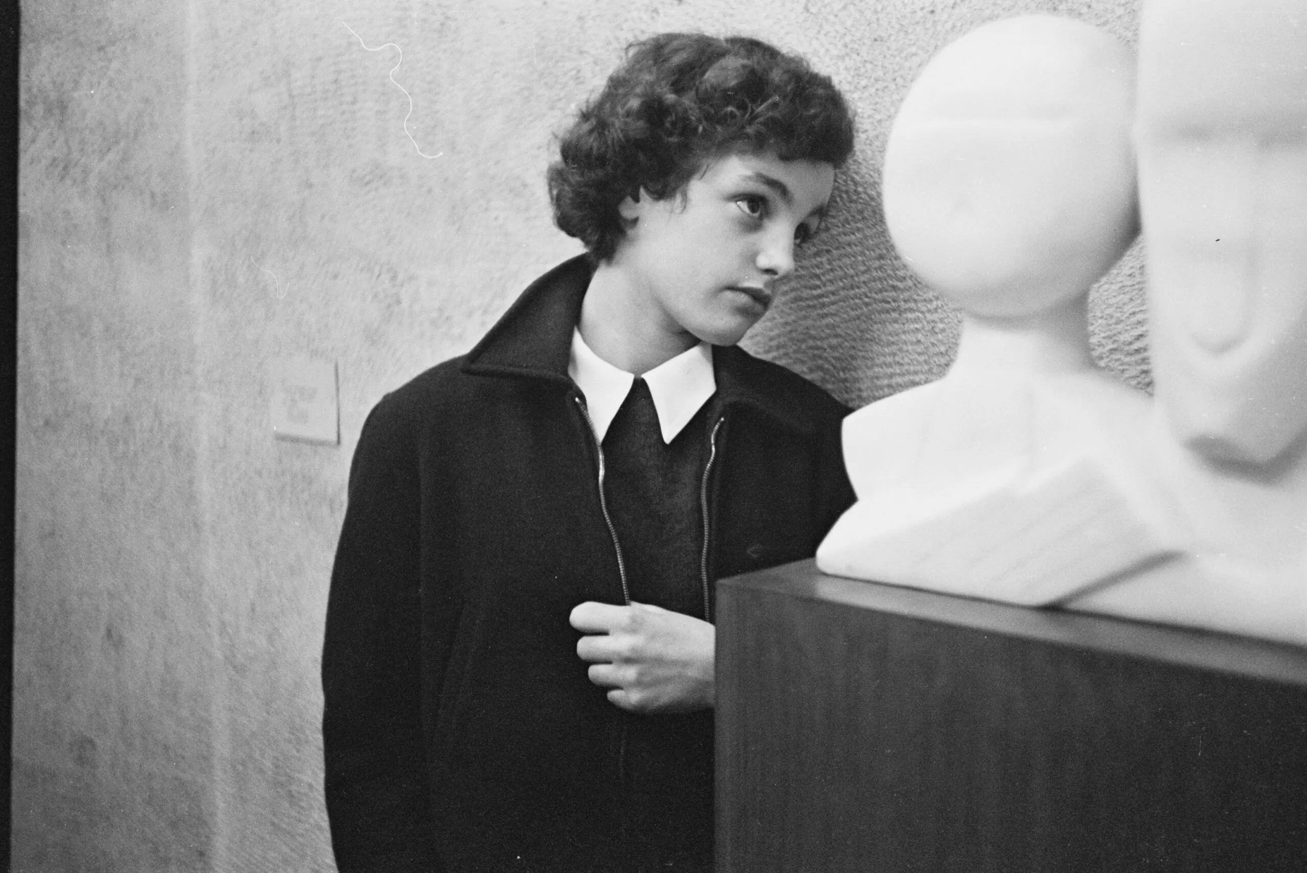 black and white photograph of a child in a dark zip jacket and white collar leaning against a textured wall, looking at a white sculpted head on a pedestal in the foreground.