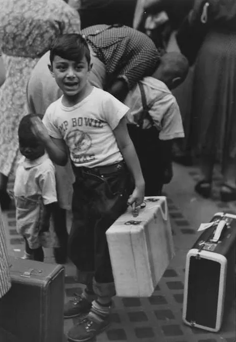 black and white photograph of a smiling boy in a camp t-shirt carrying a small suitcase, among travelers and luggage on a station platform.