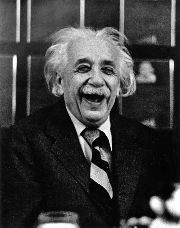 black and white photograph of Albert Einstein laughing, in a dark suit and striped tie, in front of a paned window.
