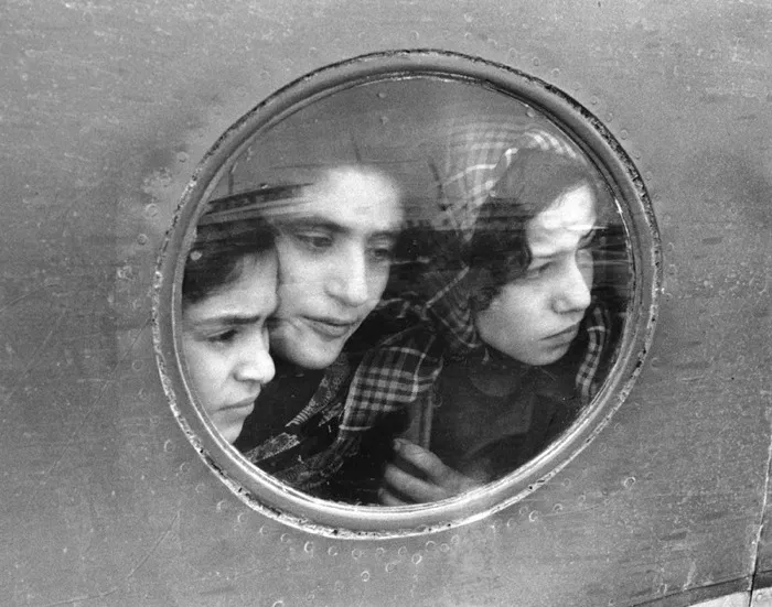 black and white photograph of three young women looking out through the round window of an airplane.