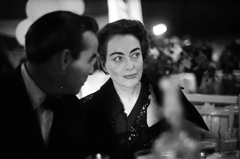 black and white photograph of Joan Crawford seated at a table at night, glancing sideways toward a man in the foreground, with lights and glassware blurred around them.