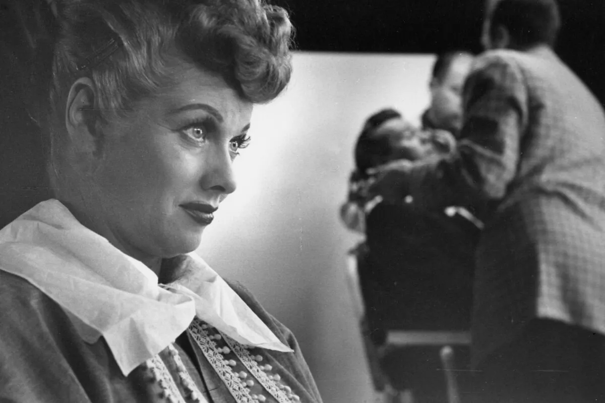 black and white photograph of Lucille Ball in profile with a tissue tucked into her collar, as crew members work in the blurred background.