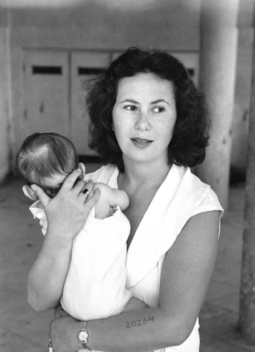 black and white photograph of a woman holding a baby against her shoulder, a number tattooed on her forearm.