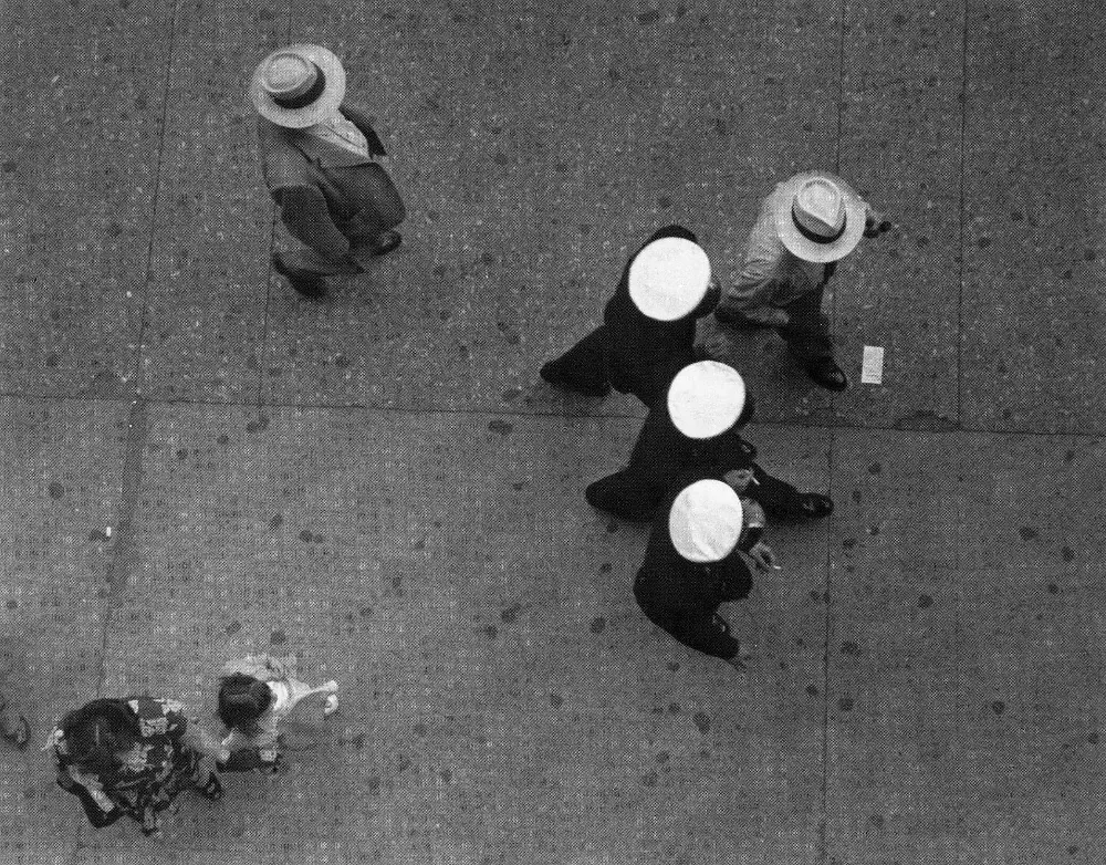 black and white photograph looking straight down at a sidewalk, where three sailors in white caps walk past two men in straw hats and a woman with a small child.