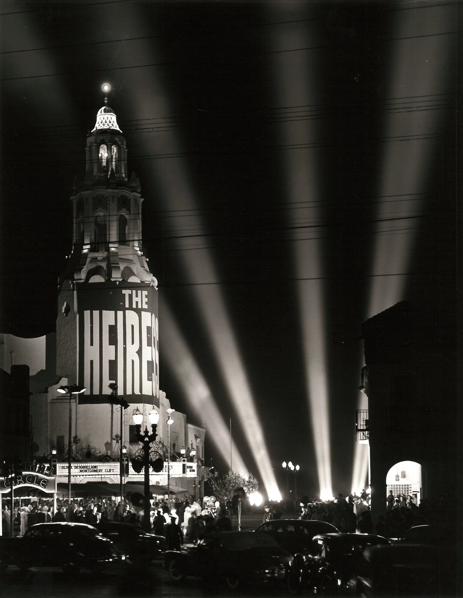 black-and-white photograph of searchlight beams fanning across the night sky over the Carthay Circle Theatre, its tower hung with a banner for The Heiress, with crowds and cars on the street below.