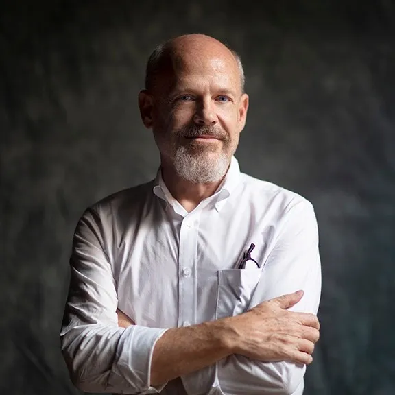 color photograph of Randy West in a white button-down shirt with his arms crossed, against a dark mottled backdrop.