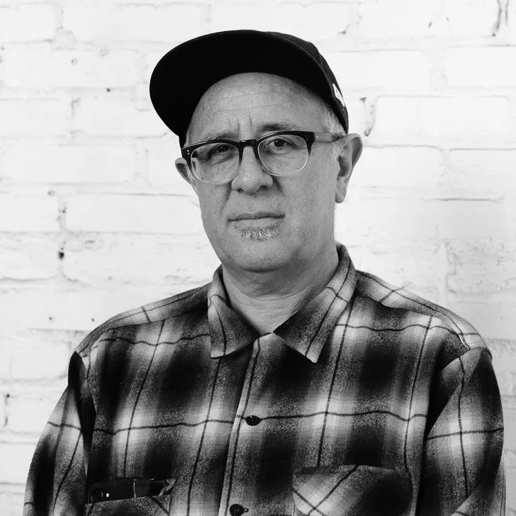 black and white photograph of Rubén Ortiz Torres in glasses, a baseball cap, and a plaid flannel shirt, in front of a white brick wall.