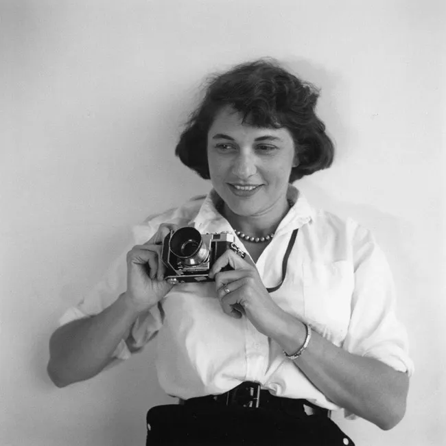 black and white photograph of Ruth Orkin smiling against a pale wall, holding a camera at her chest, in a white shirt and pearls.